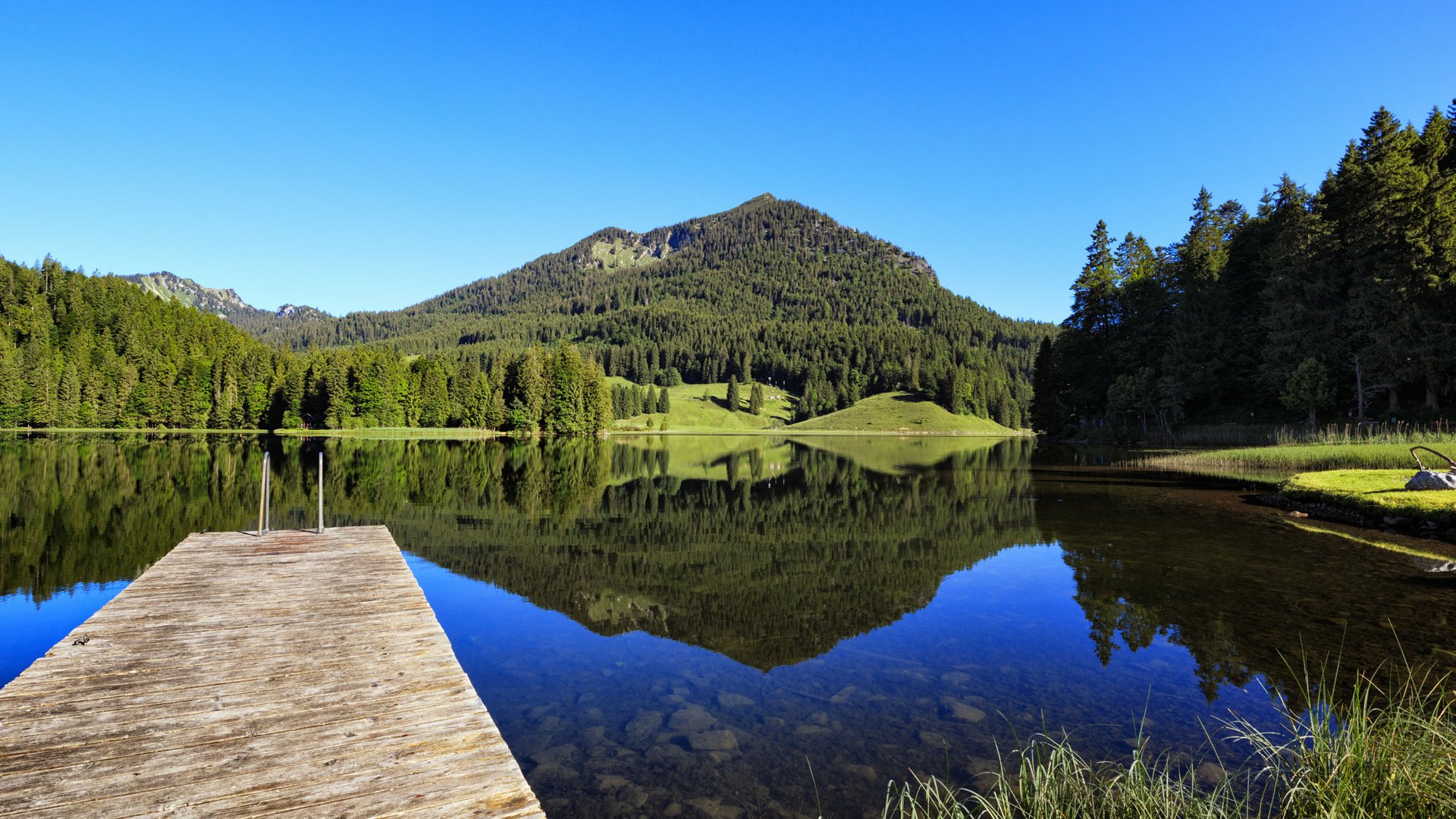 Euer authentischer Urlaub am Spitzingsee Steg an einem ruhigen Bergsee mit Wälder- und Bergreflexion bei klarem Himmel