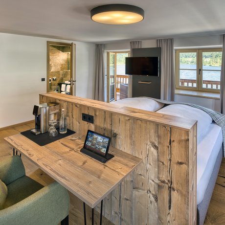 Ein Blick in die Alte Wurzhütte Helles Hotelzimmer mit Holzmöbeln, Schreibtisch, Sessel und Seeausblick