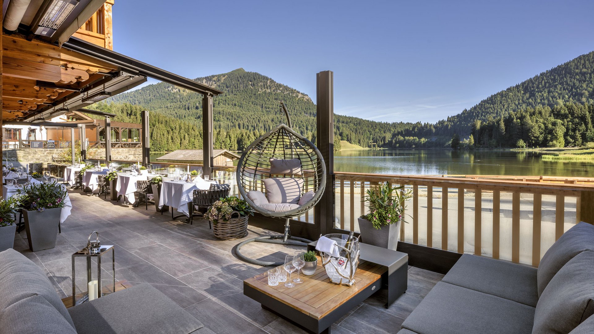 Mit einem Lächeln aufstehen im Hotel am Spitzingsee Gemütliche Terrasse mit Seeblick, Hängesessel und Tischen vor Bergkulisse