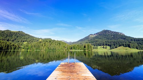 Mit einem Lächeln aufstehen im Hotel am Spitzingsee Steg führt zu ruhigem Bergsee mit Wald und Bergen im Hintergrund